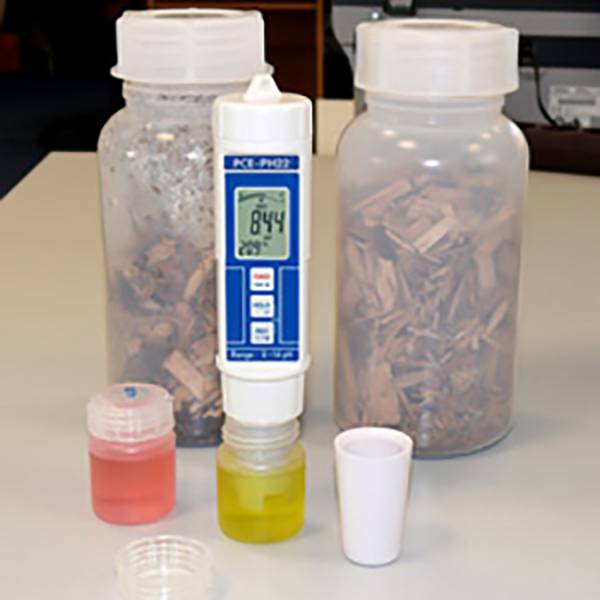 Messgerät zur Messung des pH-Werts von Holzchips, umgeben von Probenfläschchen mit Flüssigkeiten. Angezeigter Wert: 8,44.