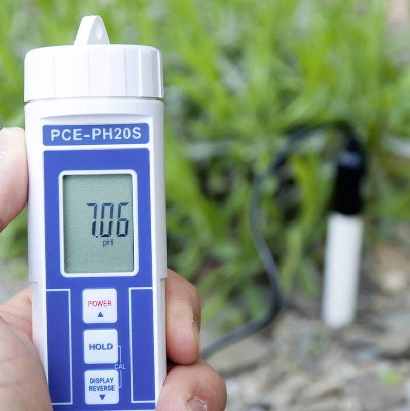 Eine Hand hält ein pH-Meter mit der Anzeige '7.06 pH'. Im Hintergrund ist Gras sichtbar.