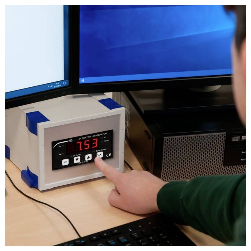 Person drückt einen Knopf auf einem digitalen Temperaturregler, auf dem 7.53 angezeigt wird, neben zwei Computermonitoren.