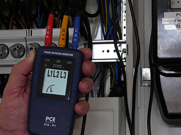 Eine Person hält einen Phasenfolge-Indikator, der die Phasen L1, L2 und L3 anzeigt. Hintergrund zeigt ein offenes Schaltschranksystem.