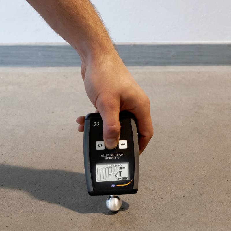 Hand hält ein Messgerät zur Feuchtigkeitsmessung auf einem Betonboden. Das Display zeigt einen Balkenwert an.