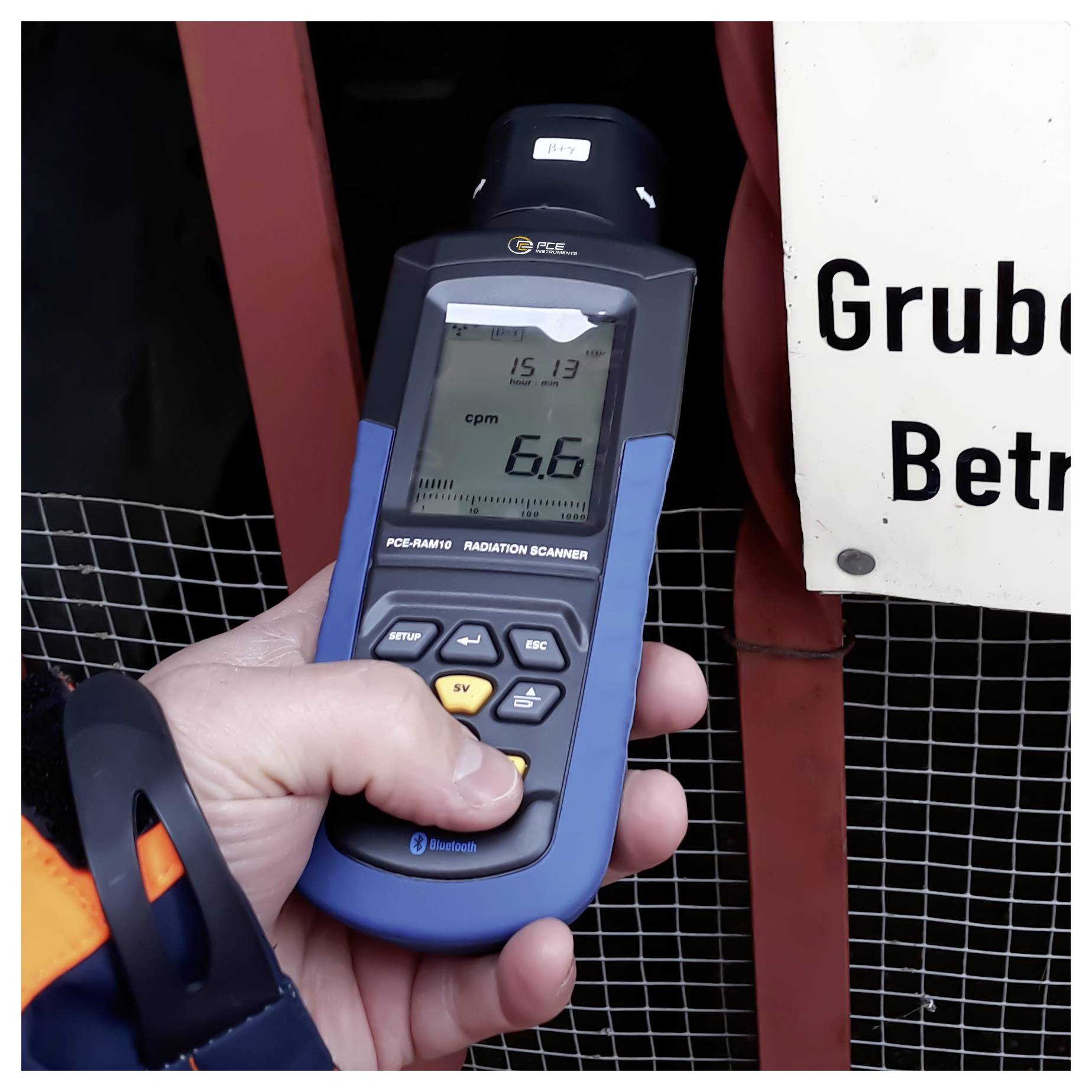 Tragbarer Strahlungsdetektor, der 6,6 cpm anzeigt, gehalten in der Nähe einer Barriere mit einem sichtbaren Warnschild 