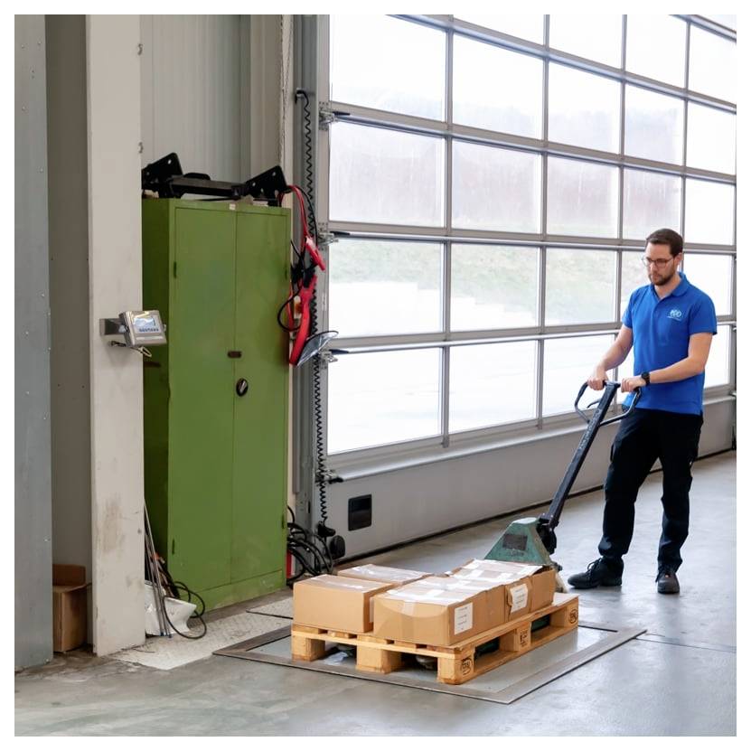 Arbeiter in blauem Hemd, der einen Hubwagen verwendet, um eine Palette mit verpackten Kartons in einem Lagerhaus oder Ladebereich zu bewegen.