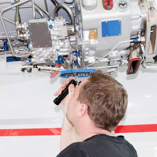 Ein Techniker inspiziert mit einer Taschenlampe den komplexen Maschinenteil eines Flugzeugmotors von unten.