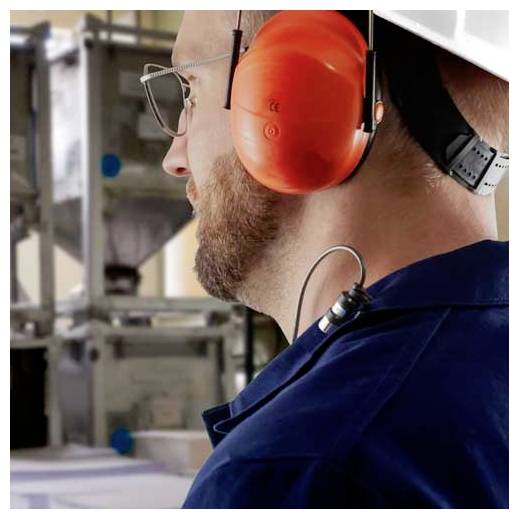 Seitenansicht eines Arbeiters, der orange Gehörschutz und Schutzbrille in einer Industrieumgebung trägt und auf Maschinen schaut.