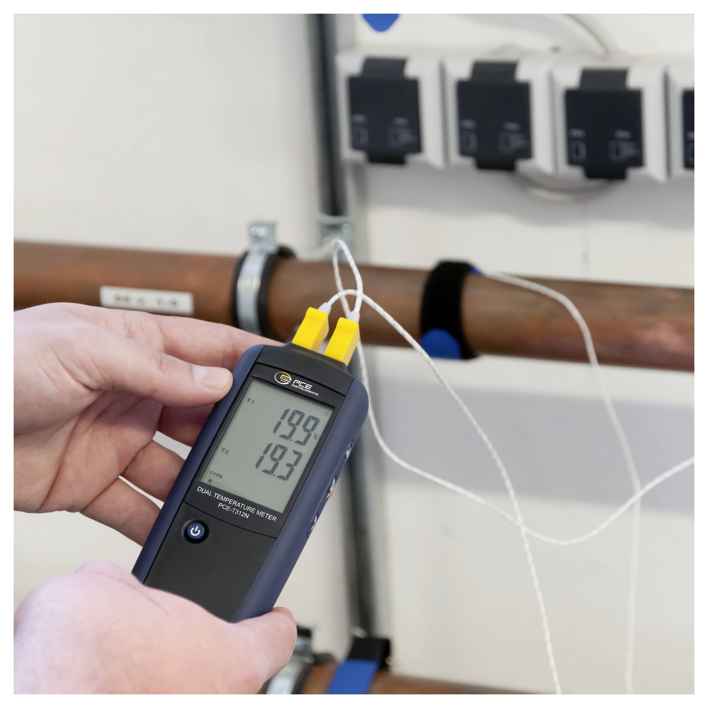 Eine Hand hält ein digitales Thermometer mit zwei Messfühlern gegen ein Rohr und zeigt Messwerte von etwa 19,9 und 19,3 Grad an.