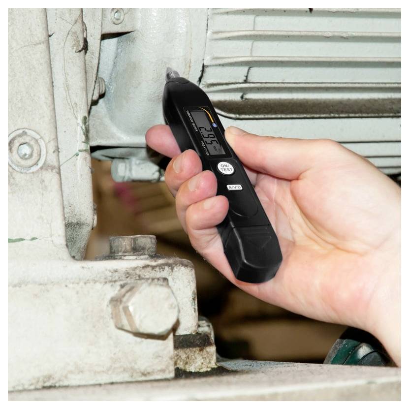 Hand, die ein digitales Infrarot-Thermometer neben einem Metallteil einer Maschine hält und die Oberflächentemperatur misst.