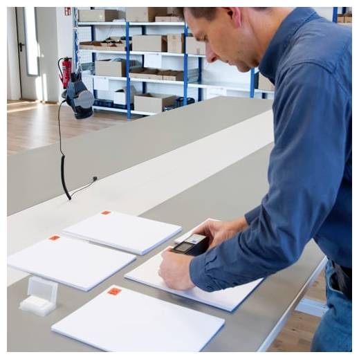 Mann im blauen Hemd, der ein weißes Paneel auf einer Werkbank in einer Werkstatt mit einem tragbaren Messinstrument misst, während sich weitere Paneele in der Nähe befinden.