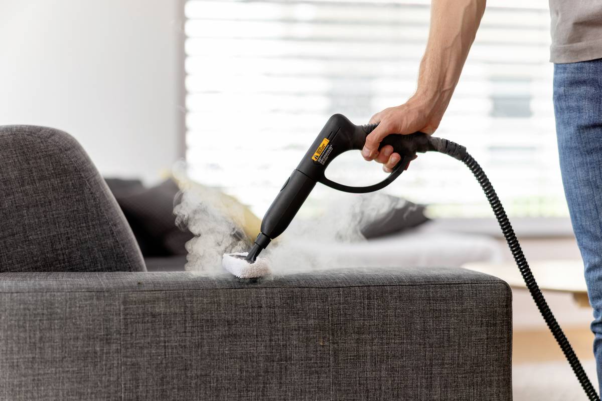 Eine Person reinigt ein graues Sofa mit einem Dampfreiniger, aus dem Dampf austritt. Im Hintergrund ist ein Fenster mit Jalousien.