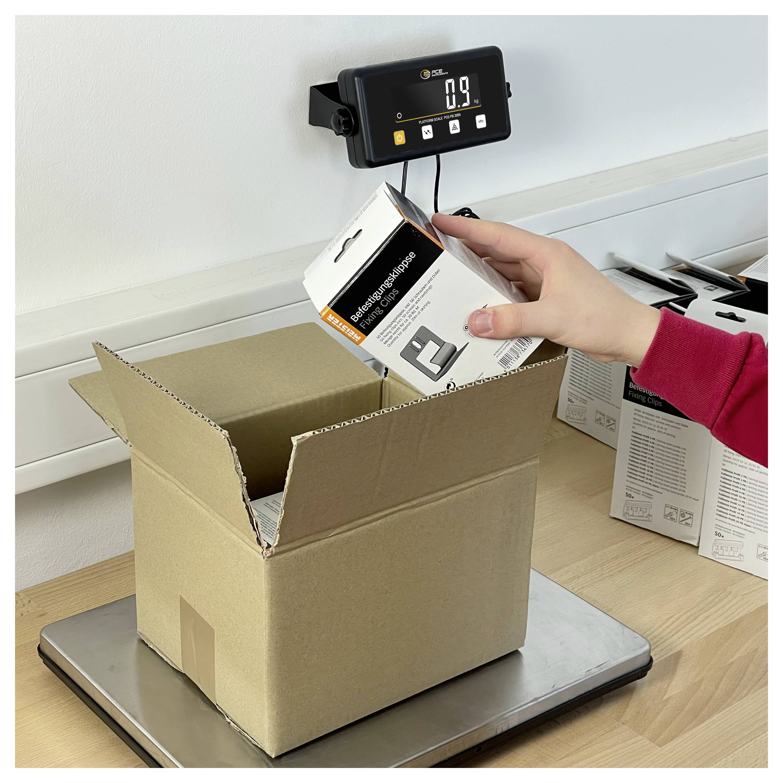 Eine Person legt eine Einzelhandelproduktschachtel in einen Kartonversandkarton auf einer digitalen Waage, die 0,9 kg anzeigt.