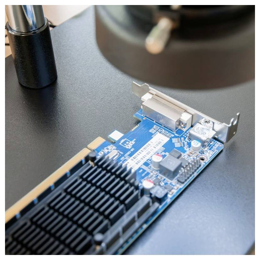 Nahaufnahme einer blauen Computererweiterungskarte mit großem schwarzem Kühlkörper und Metallhalterung auf einem Schreibtisch unter einem Mikroskop oder Inspektionswerkzeug.