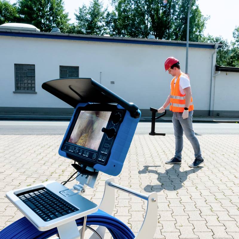 Ein Techniker mit Schutzmütze und Warnweste bedient ein tragbares Inspektionsgerät auf einer Pflasterfläche vor einem Gebäude.