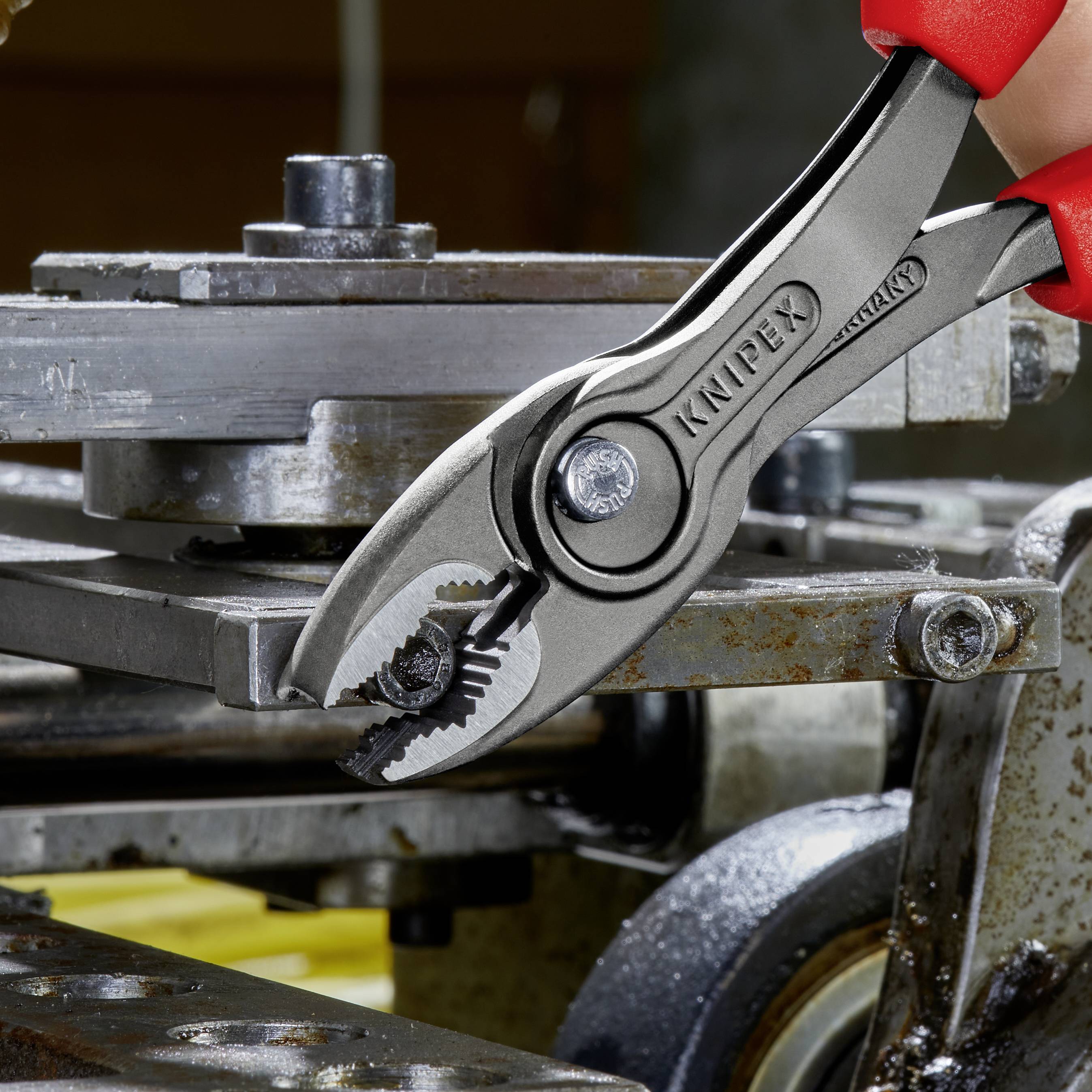Rote Hand hält Knipex-Zange, die ein Stück Metall in einer Werkstatt greift. Im Hintergrund sind industrielle Geräte zu sehen.