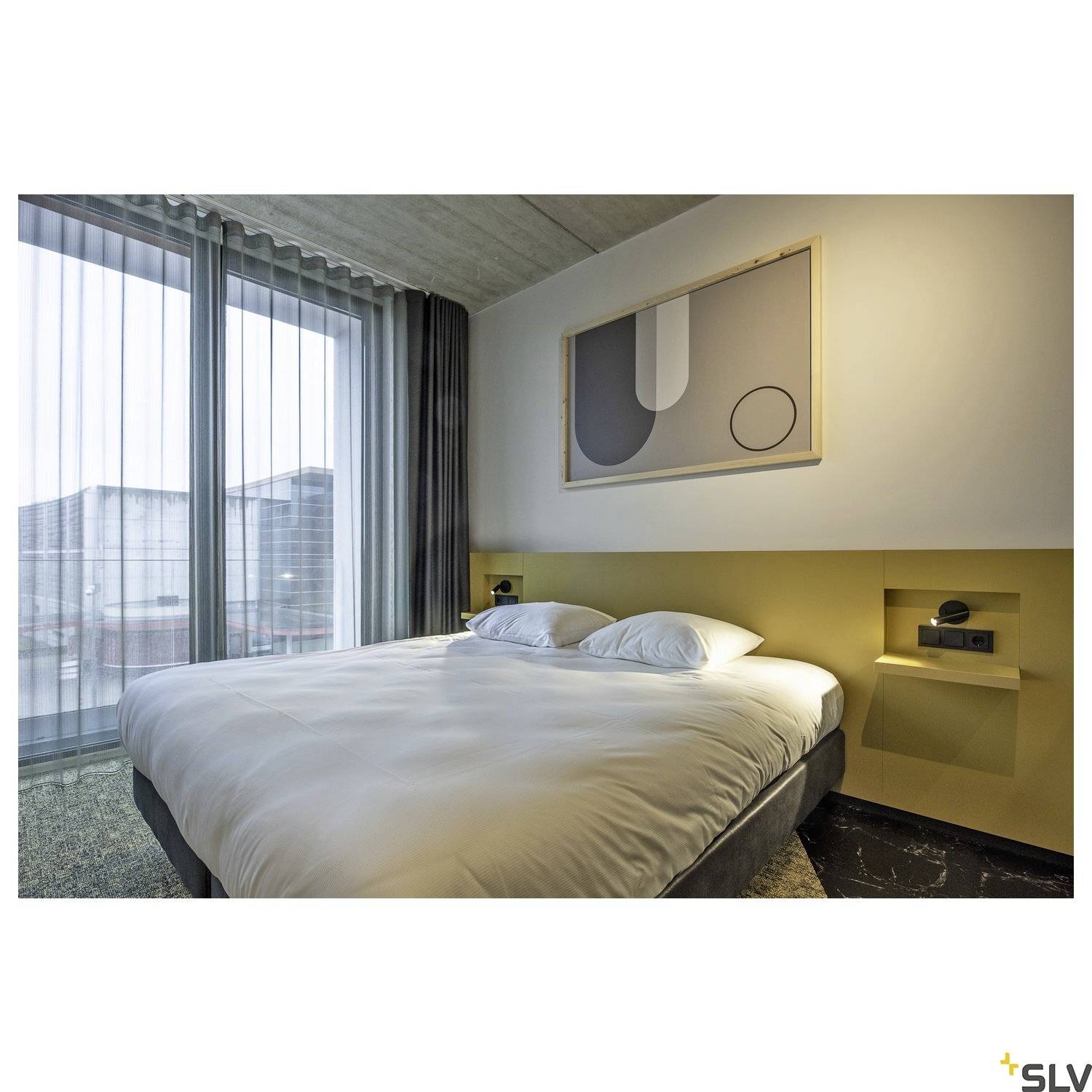 Helles Hotelzimmer mit Doppelbett, modernem abstraktem Kunstwerk an der Wand, großen Fenstern mit Sicht auf Gebäude außen.