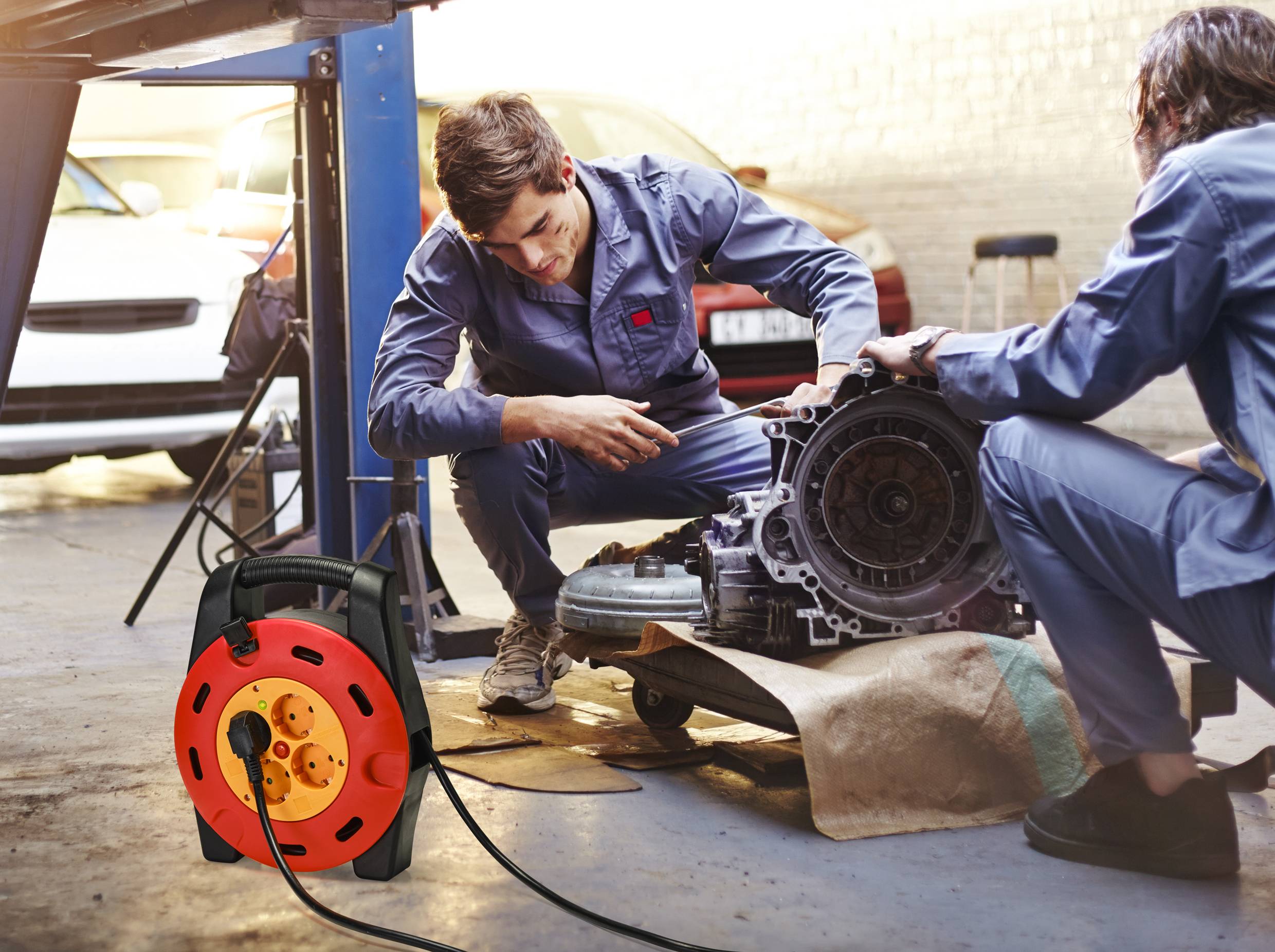 Zwei Personen in Arbeitskleidung reparieren ein großes Autoteil in einer Werkstatt. Im Vordergrund liegt eine rote Kabeltrommel.