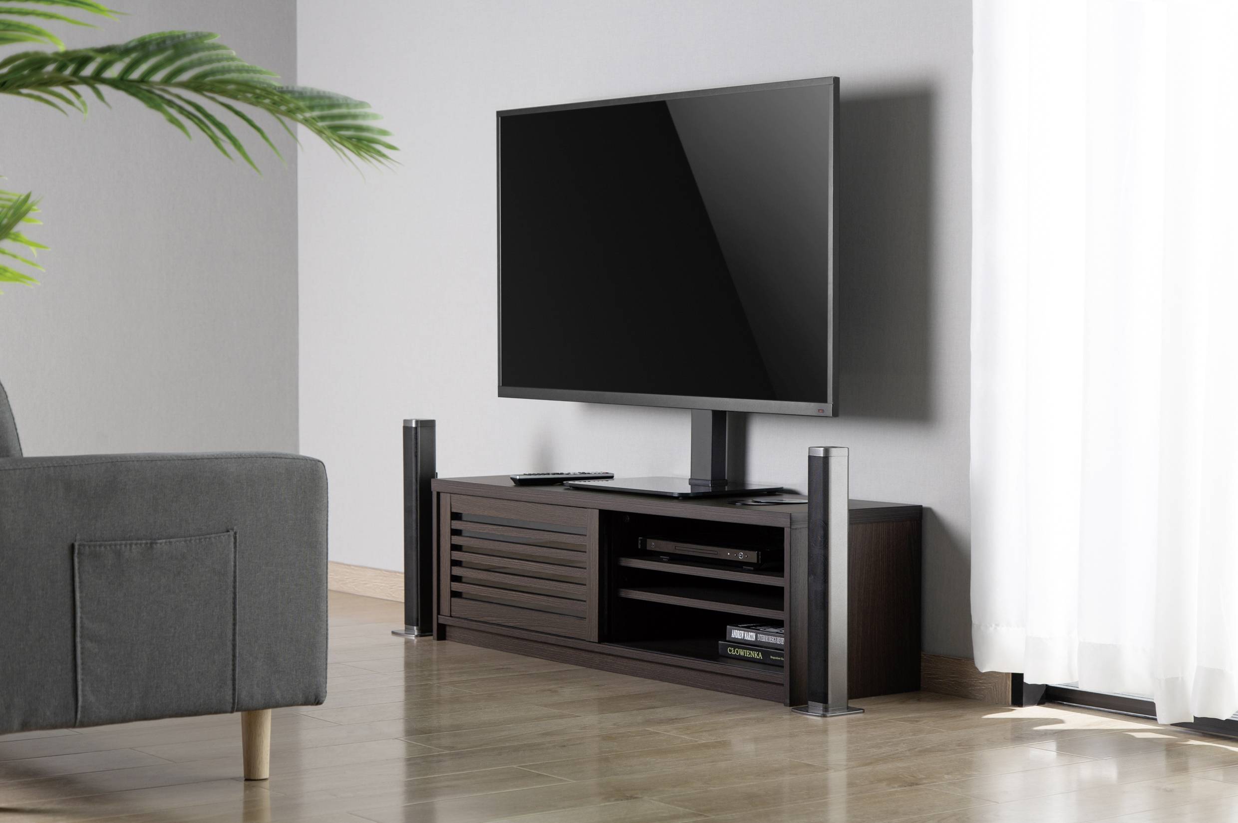 Ein moderner Fernseher steht auf einem dunklen TV-Schrank in einem hellen Wohnzimmer. Links ein graues Sofa, rechts ein Vorhang.