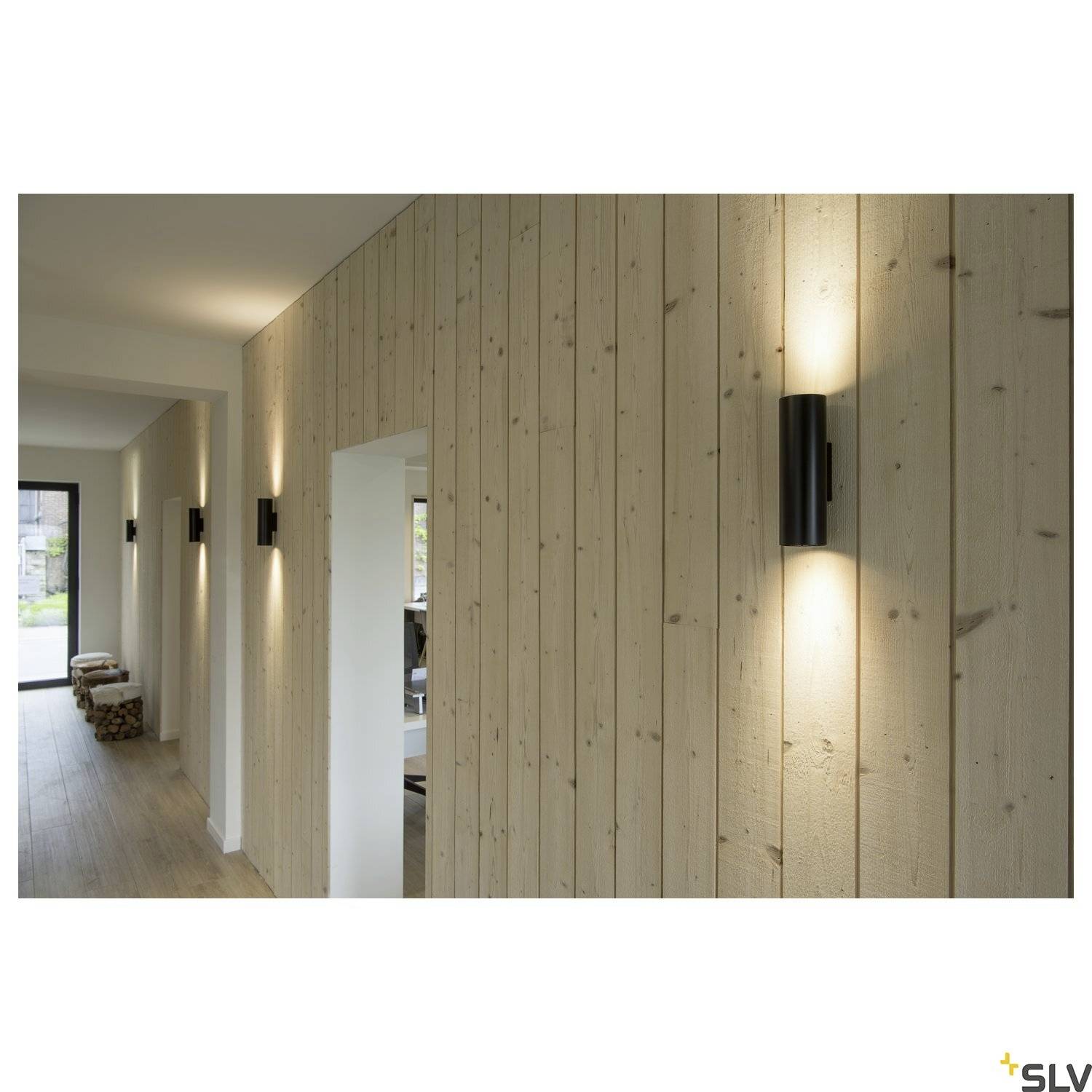 Ein Holzflur mit modernen schwarzen Wandleuchten, die warmes Licht auf die vertäfelte Wand werfen, neben einem offenen Durchgang.