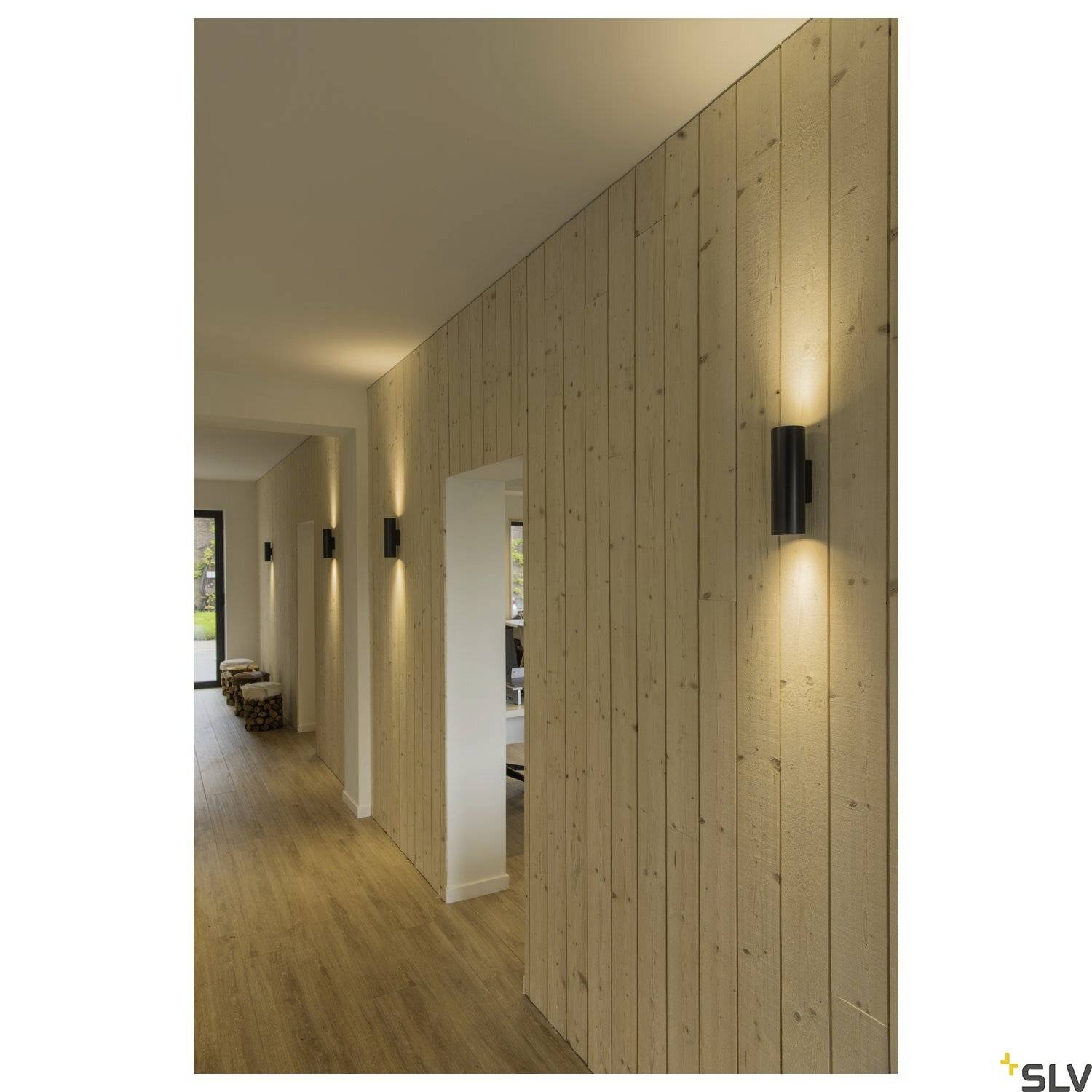 Korridor mit Holzverkleidung und modernen Wandleuchten, die ein warmes Licht verbreiten. Hölzerner Boden und mehrere Sitzgelegenheiten am Ende.