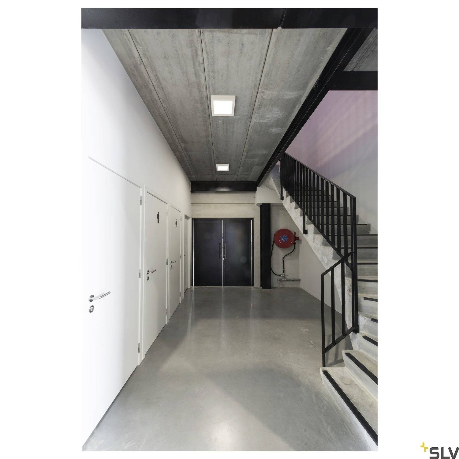 Flur mit Betonboden, Türen rechts und links, Stahltreppe mit Geländer rechts, Ausgang am Ende. Moderne, minimalistische Architektur.