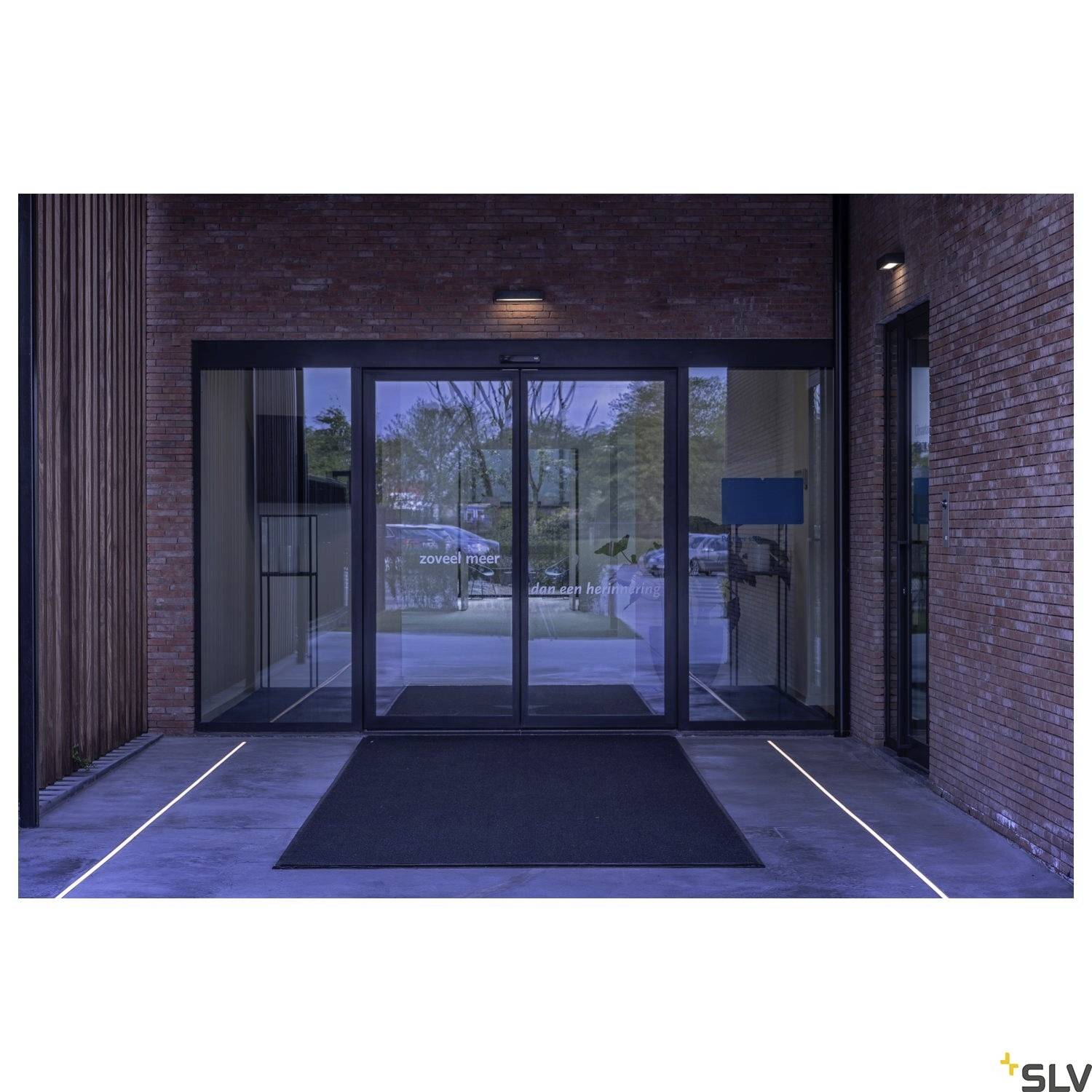 Eingang eines Gebäudes mit automatischen Glastüren, flankiert von stimmungsvoller Beleuchtung. Darauf sind Schriftzüge schwer erkennbar.