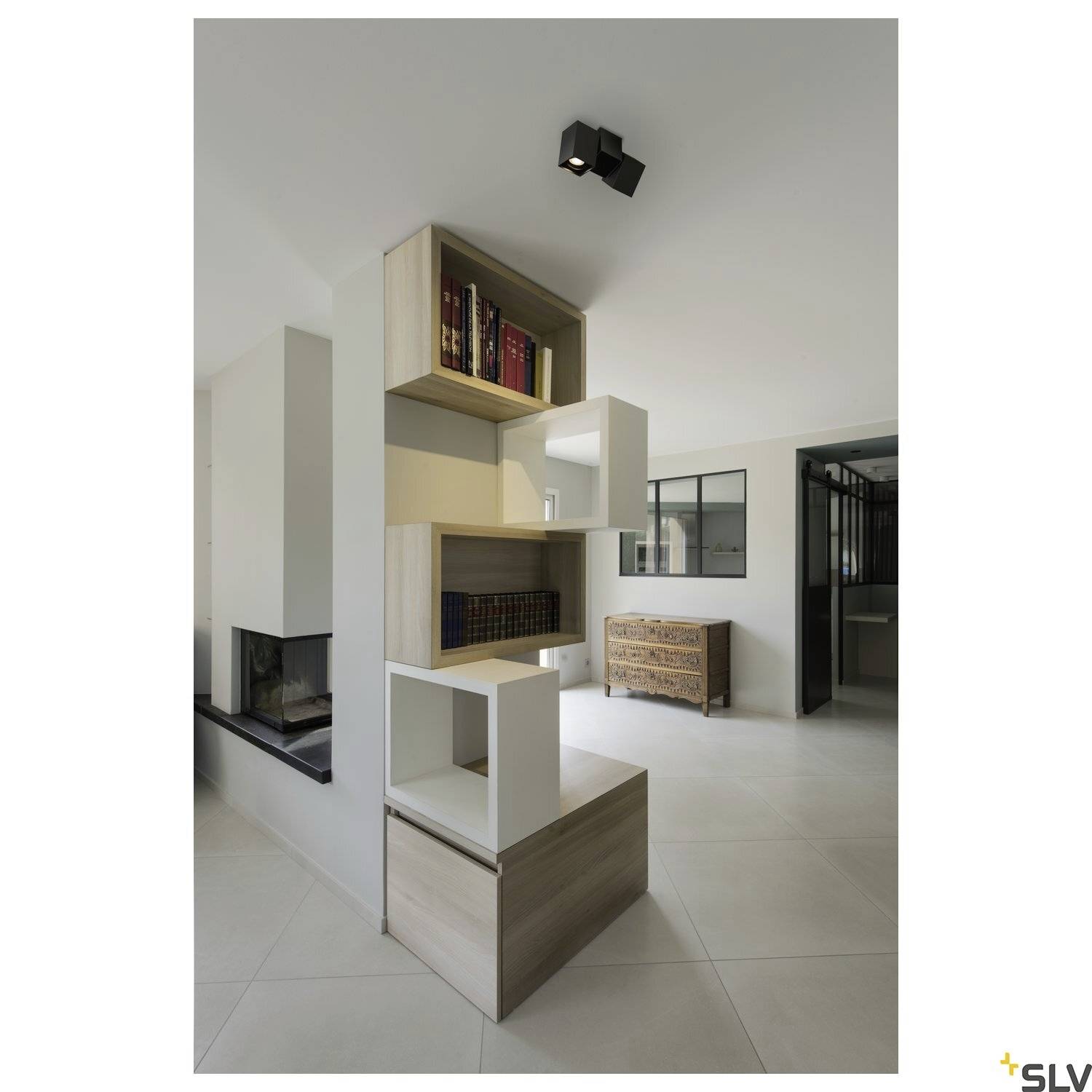 Modernes Wohnzimmer mit minimalistischem Regal, das Bücher und Dekorationen präsentiert. Helle Wände und Fliesenboden verstärken die schlichte Atmosphäre.