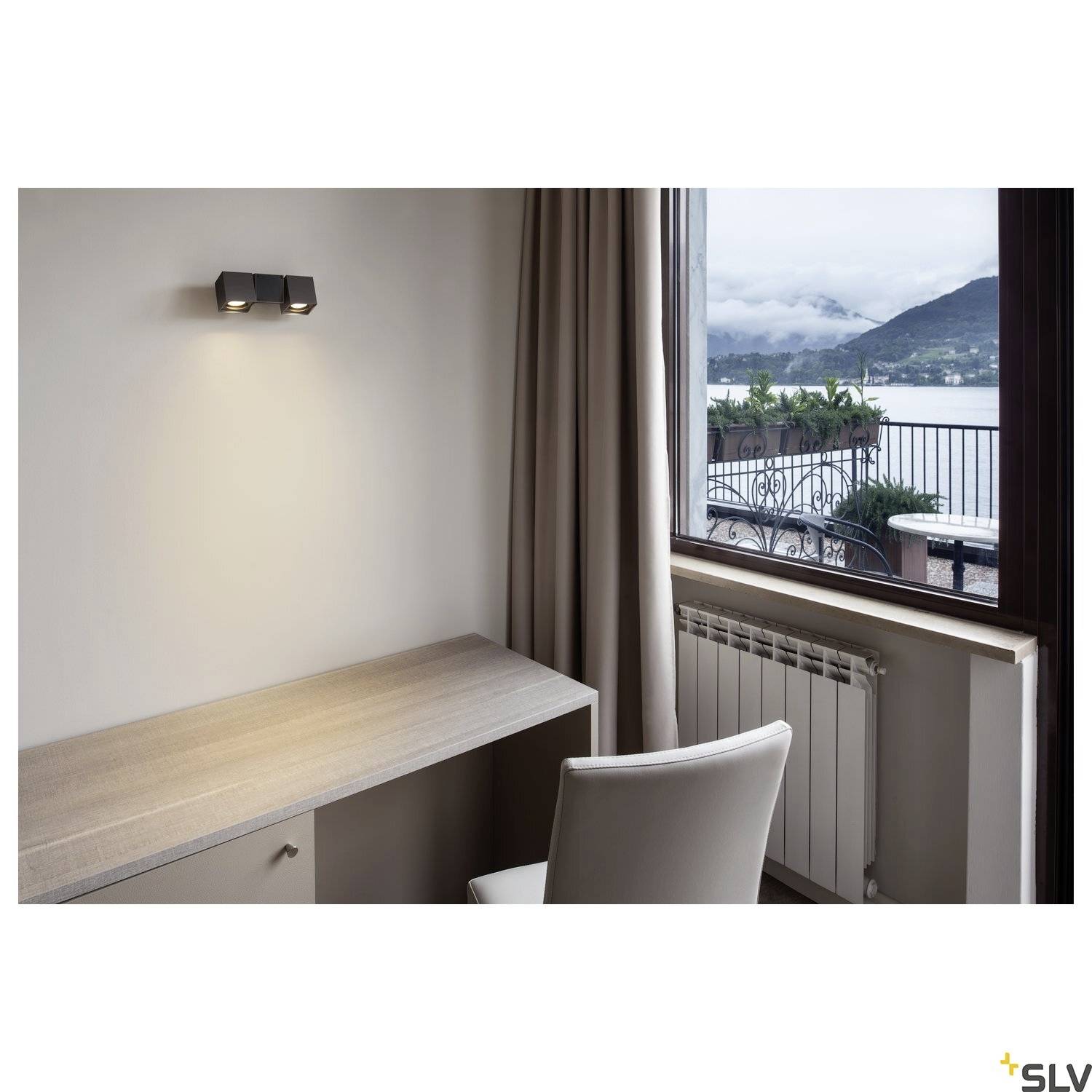 Arbeitsplatz mit Schreibtisch und Stuhl vor einem Fenster mit Blick auf einen See und Berge, Wandlicht beleuchtet den Bereich.