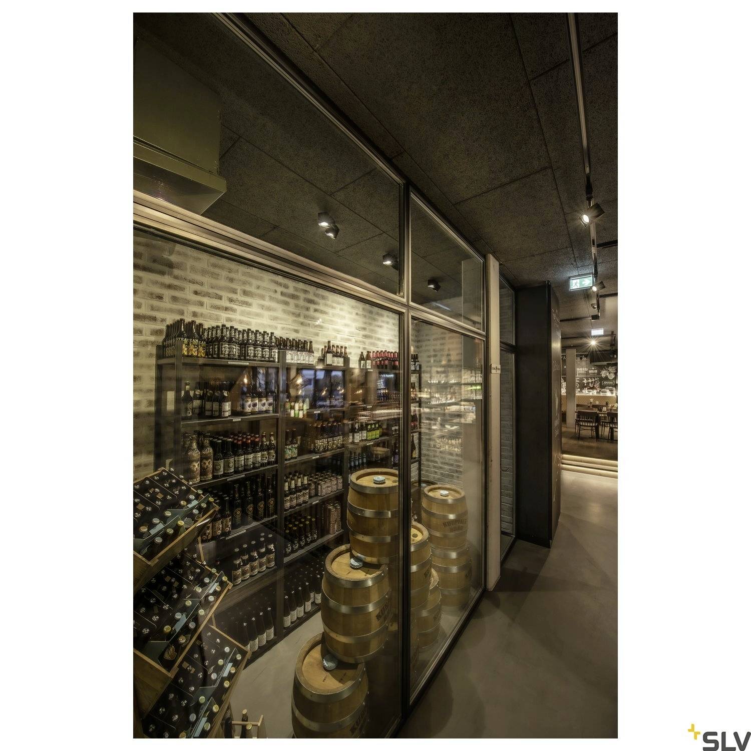 Ein Geschäftslokal zeigt Holzfässer und Regale mit Bierflaschen hinter einer Glaswand. Die Szene ist warm und gemütlich beleuchtet.