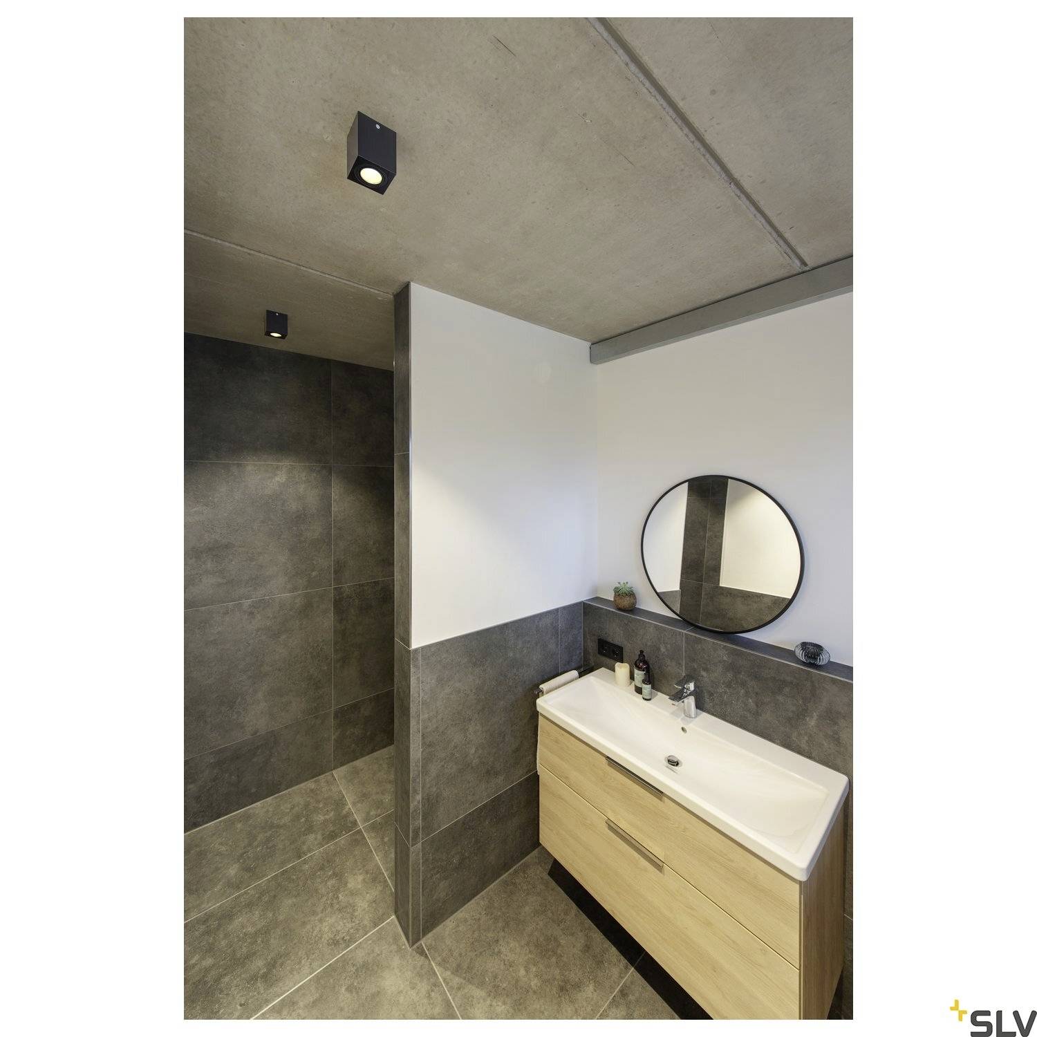 Modernes Badezimmer mit einer dunklen, gefliesten Duschwand, einem Holzwaschtisch mit zwei Waschbecken und einem runden Spiegel.