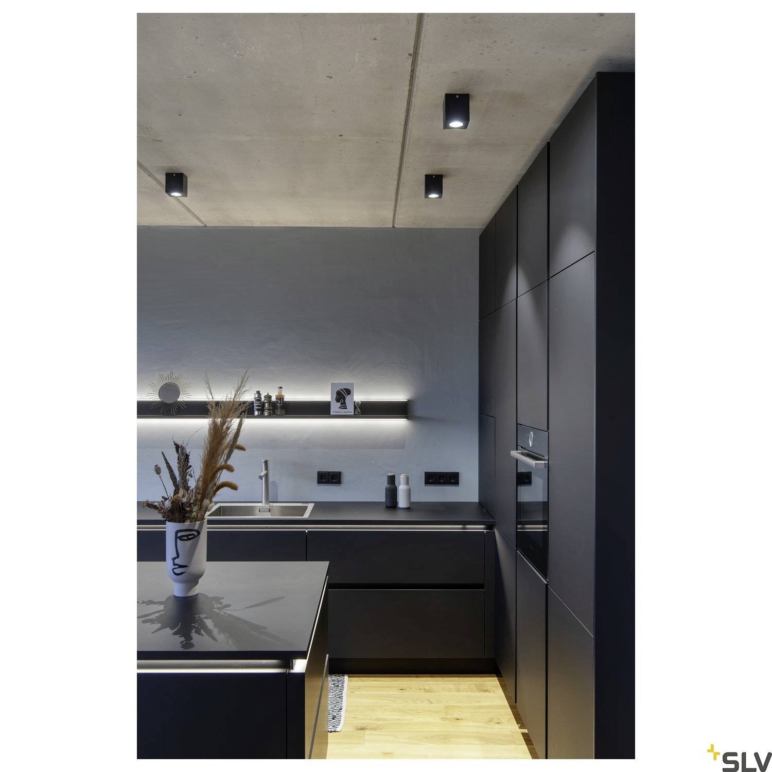 Moderne Küche mit dunklen Schränken und eingelassener Beleuchtung. Auf der Arbeitsplatte steht eine Vase mit Trockenblumen.