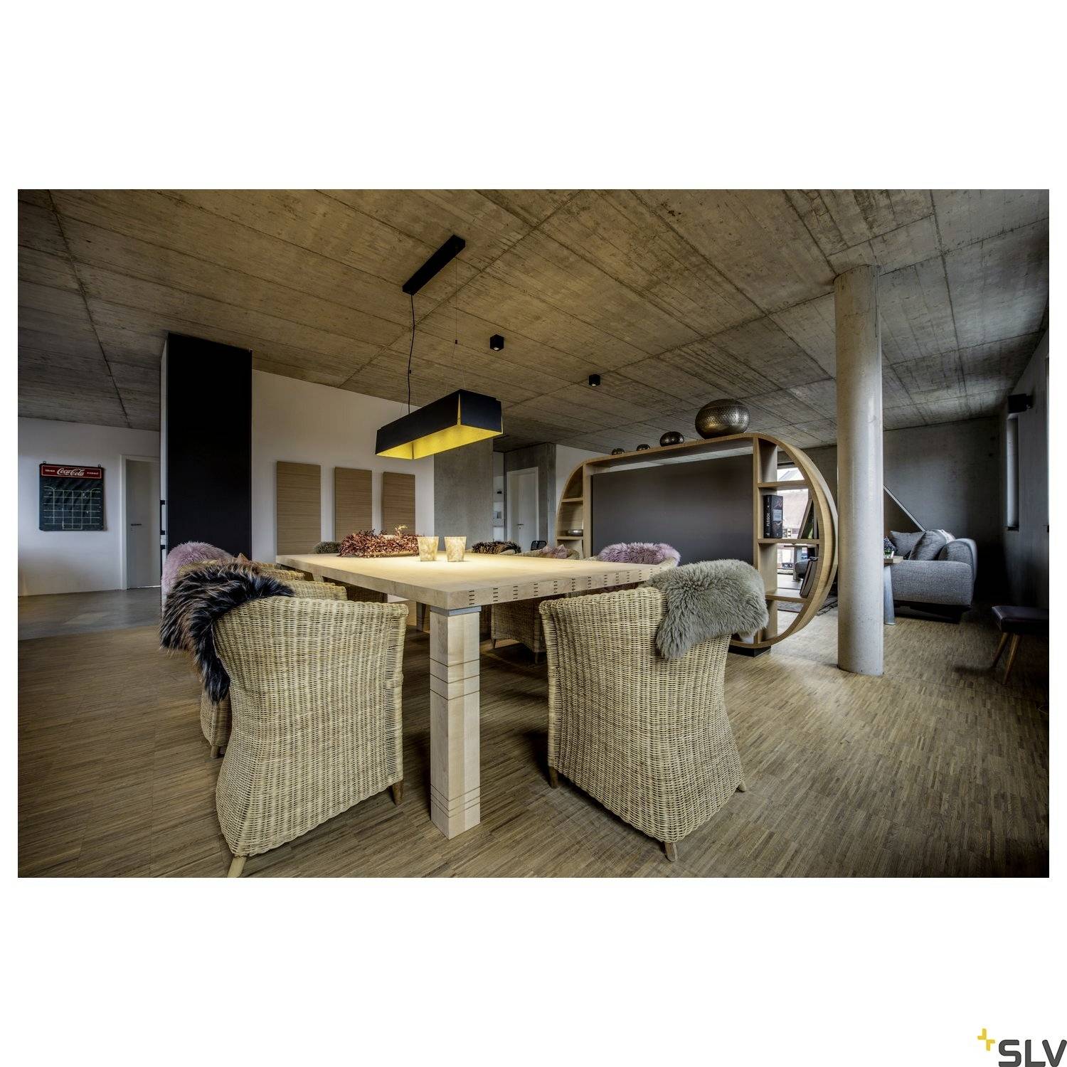 Modernes Esszimmer mit Holztisch, Korbstühlen und hängender Lampe in einem Raum mit Betonwänden und -decke. Im Hintergrund ein Sofa.