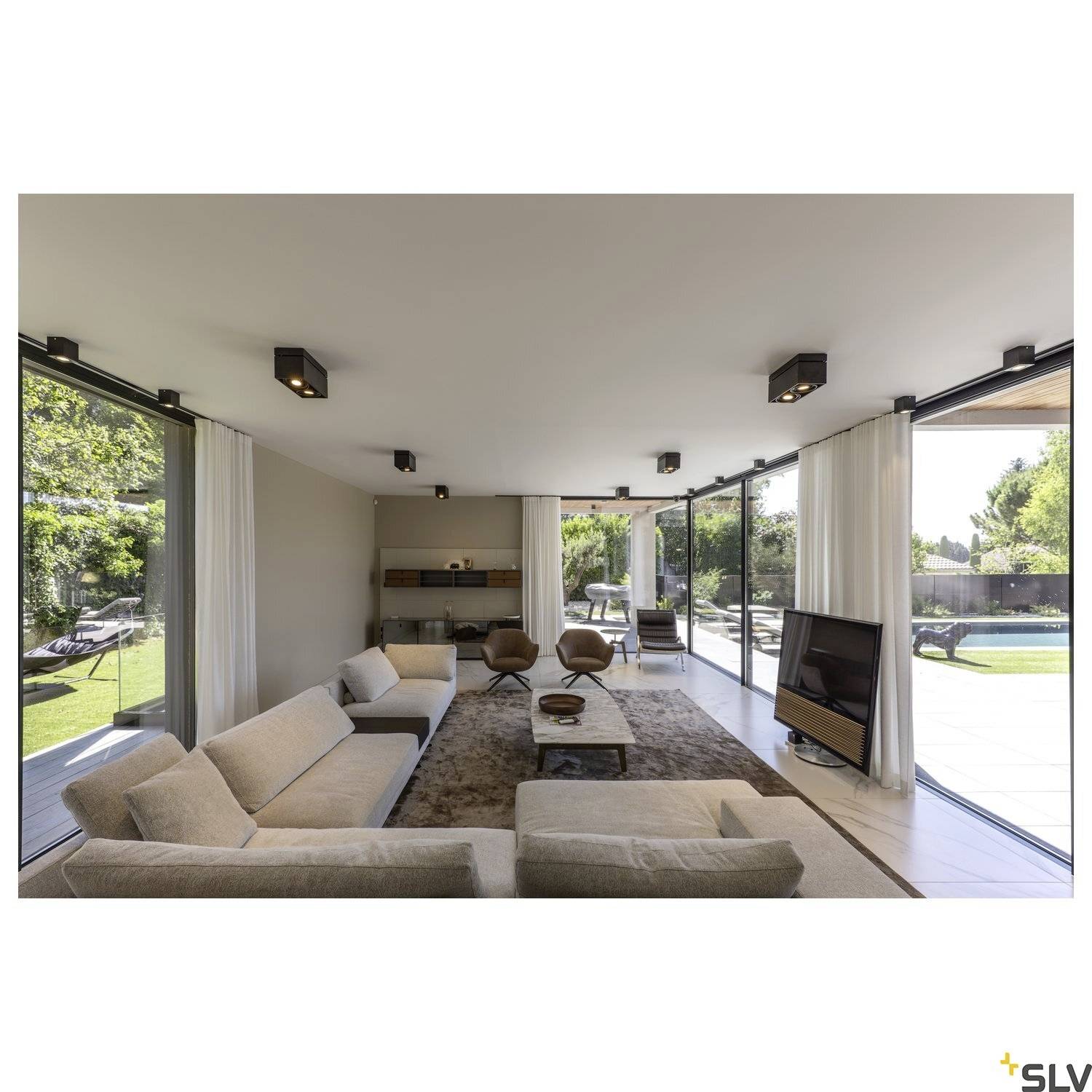 Geräumiges Wohnzimmer mit großen Glasfenstern, modernem Sofa, Teppich, TV und Blick auf einen grünen Garten. Helles und offenes Ambiente.