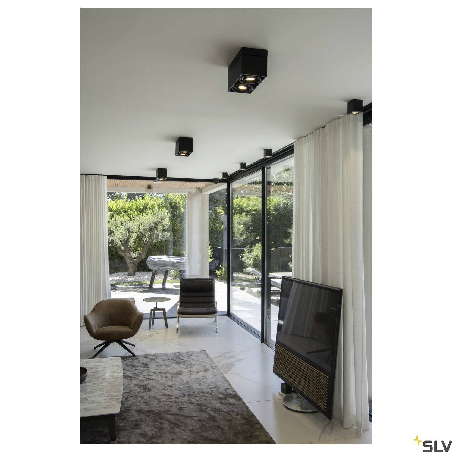 Wohnzimmer mit modernen Möbeln, großen Glasfenstern und Blick auf einen Garten. Deckenleuchten sorgen für Beleuchtung.