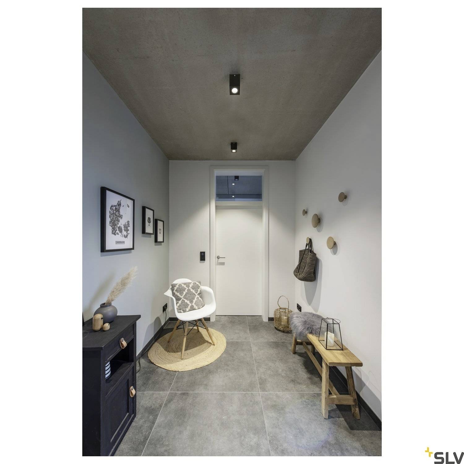 Ein minimalistischer Flur mit einer modernen Deckenleuchte. Links steht ein Stuhl auf einem kleinen runden Teppich, rechts eine Holzgarderobe.