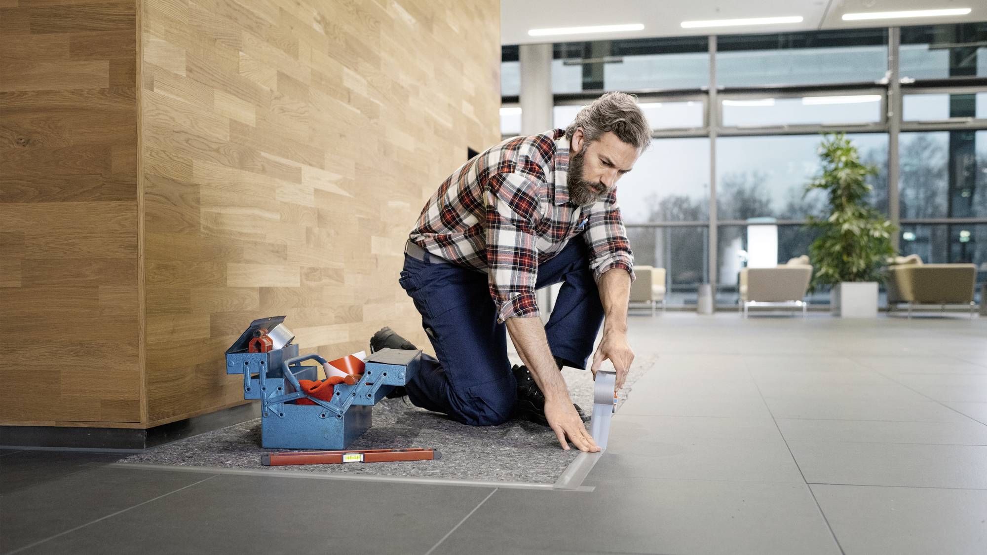 Ein Mann kniet auf einem Teppichstück, umgeben von Werkzeugen, in einem modernen Raum mit Holz- und Glaselementen.