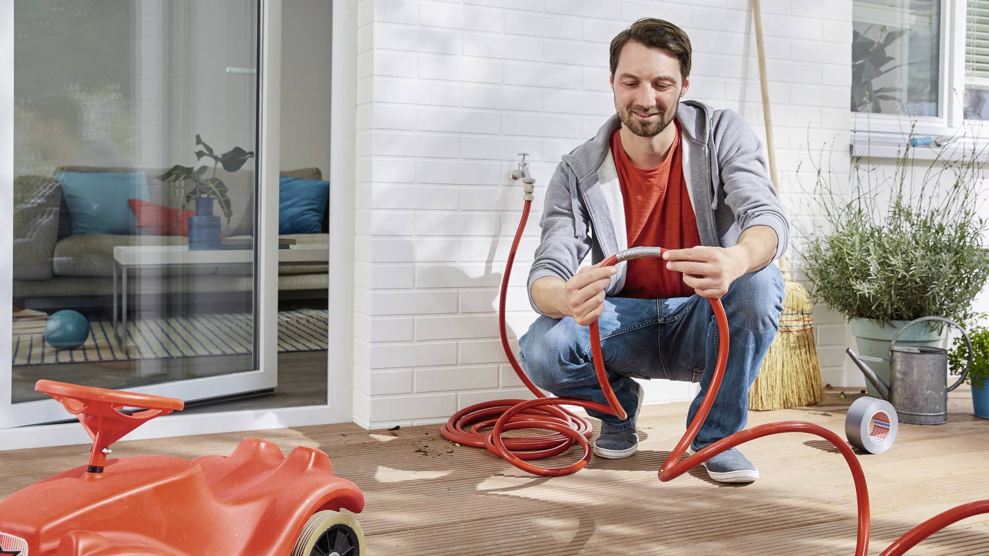 Ein Mann kniet auf einer Terrasse und repariert einen Gartenschlauch. Im Vordergrund steht ein rotes Kinderspielzeugauto.