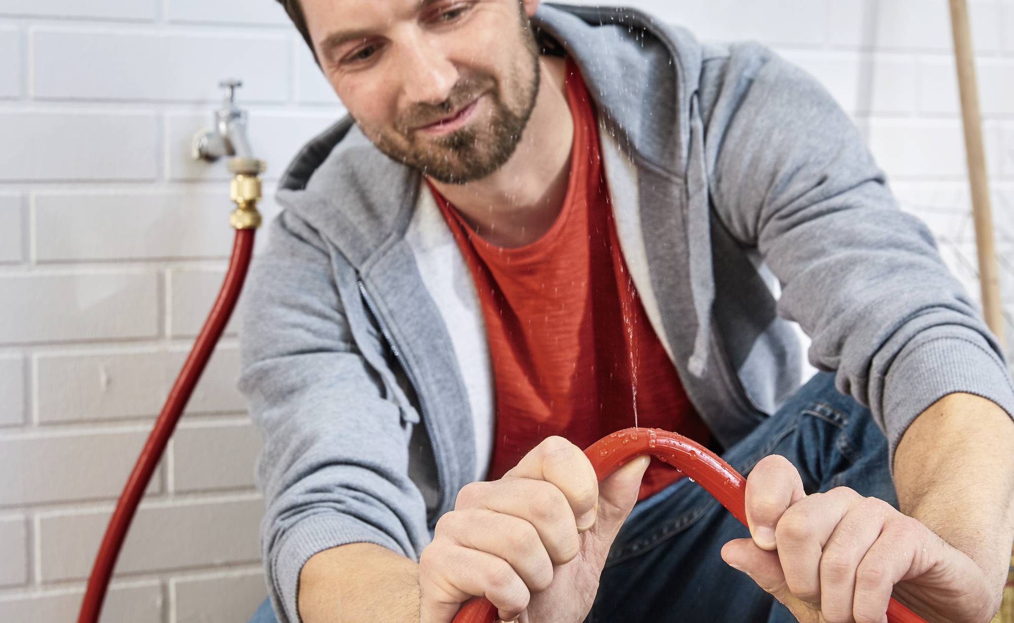 Ein Mann in einem blauen Hoodie und rotem T-Shirt befestigt einen roten Schlauch an einem Wasserhahn an einer weiß gefliesten Wand.