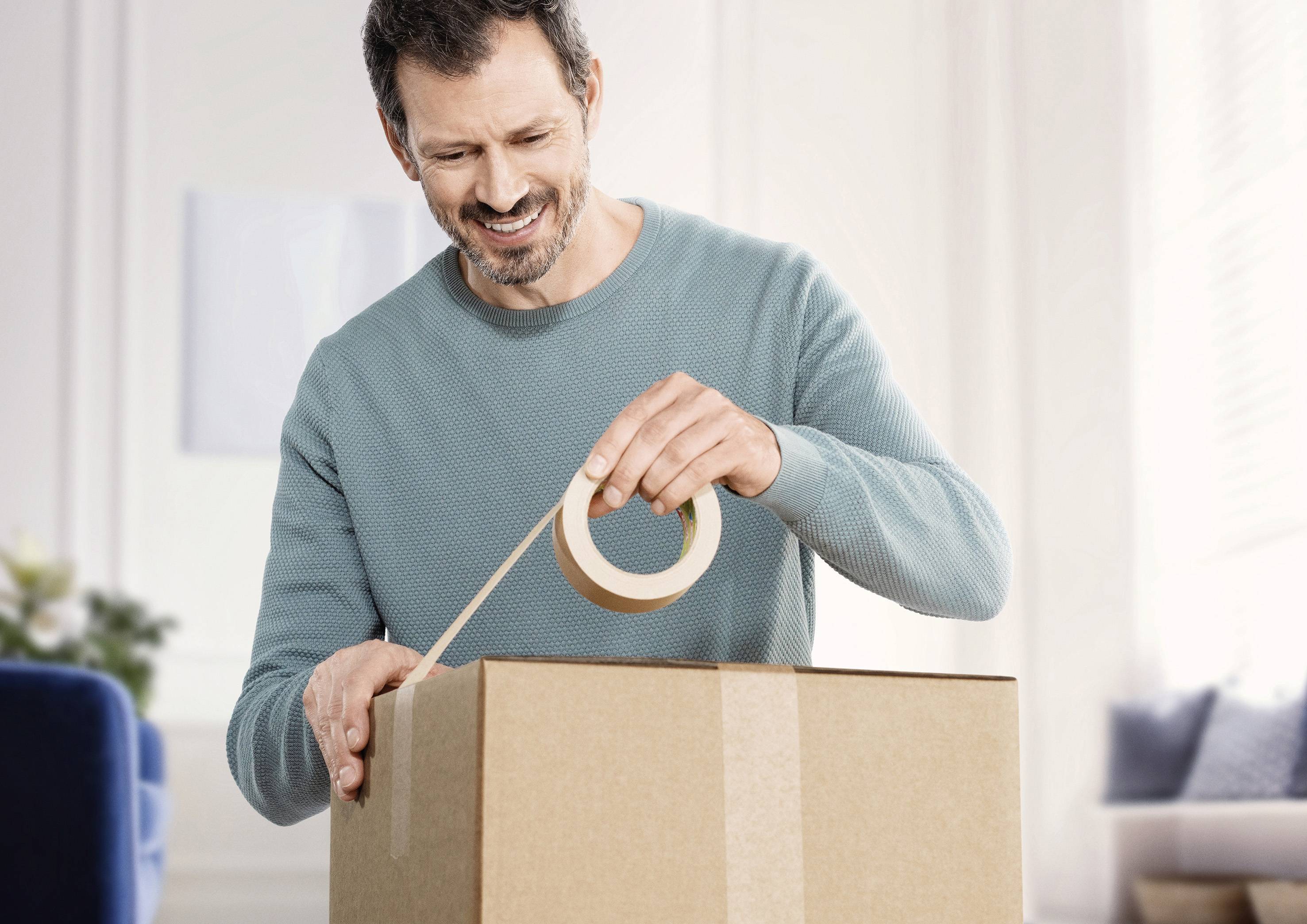 Ein Mann im blauen Pullover klebt mit Klebeband einen Karton zu. Er wirkt glücklich, das Zimmer im Hintergrund ist hell erleuchtet.