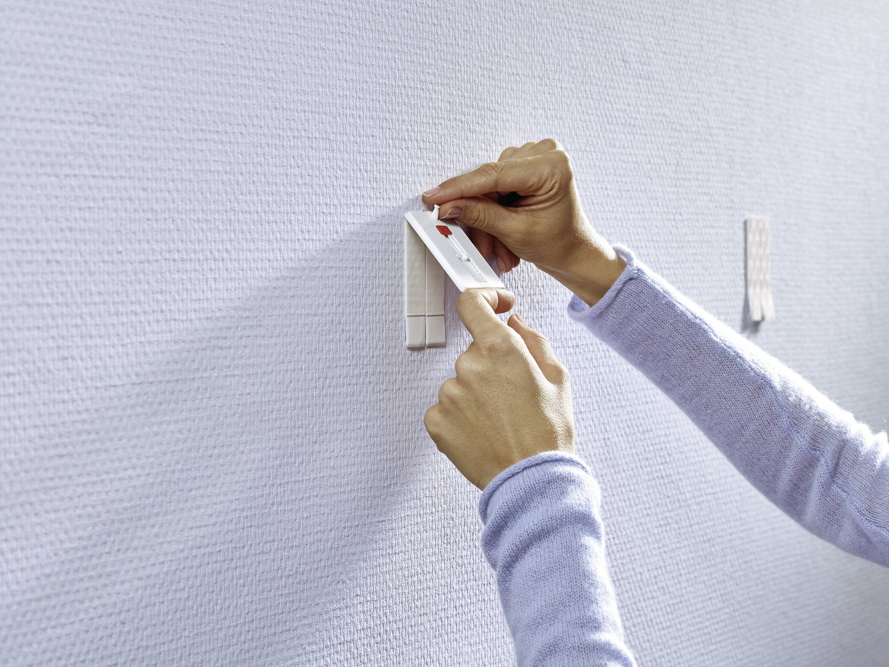 Zwei Hände befestigen mit Klebestreifen einen kleinen, rechteckigen Gegenstand an einer strukturierten, hellen Wand.