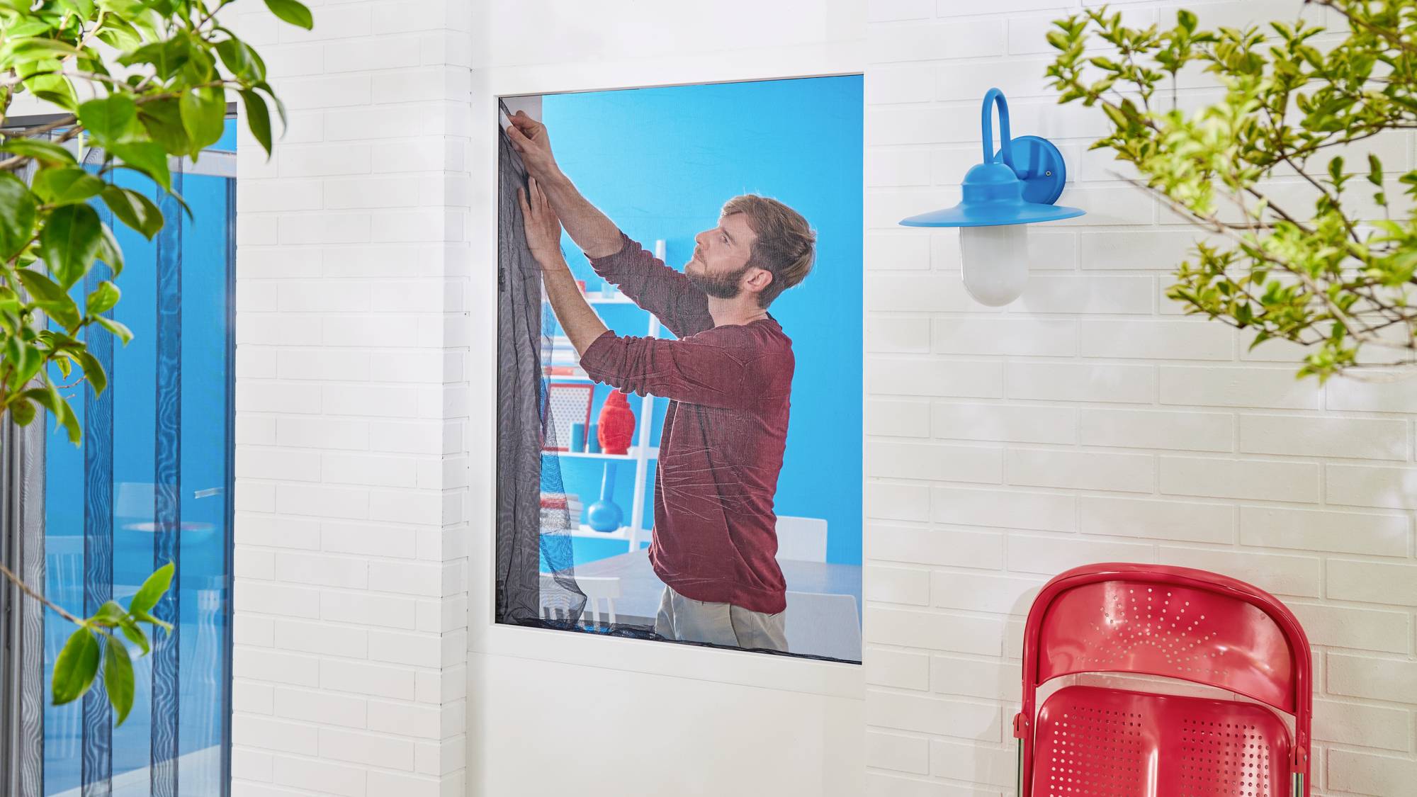 Ein Mann hängt einen Vorhang vor einem Fenster auf. Im Hintergrund sind blaue Wände, Regale und eine rote Lampe sichtbar.