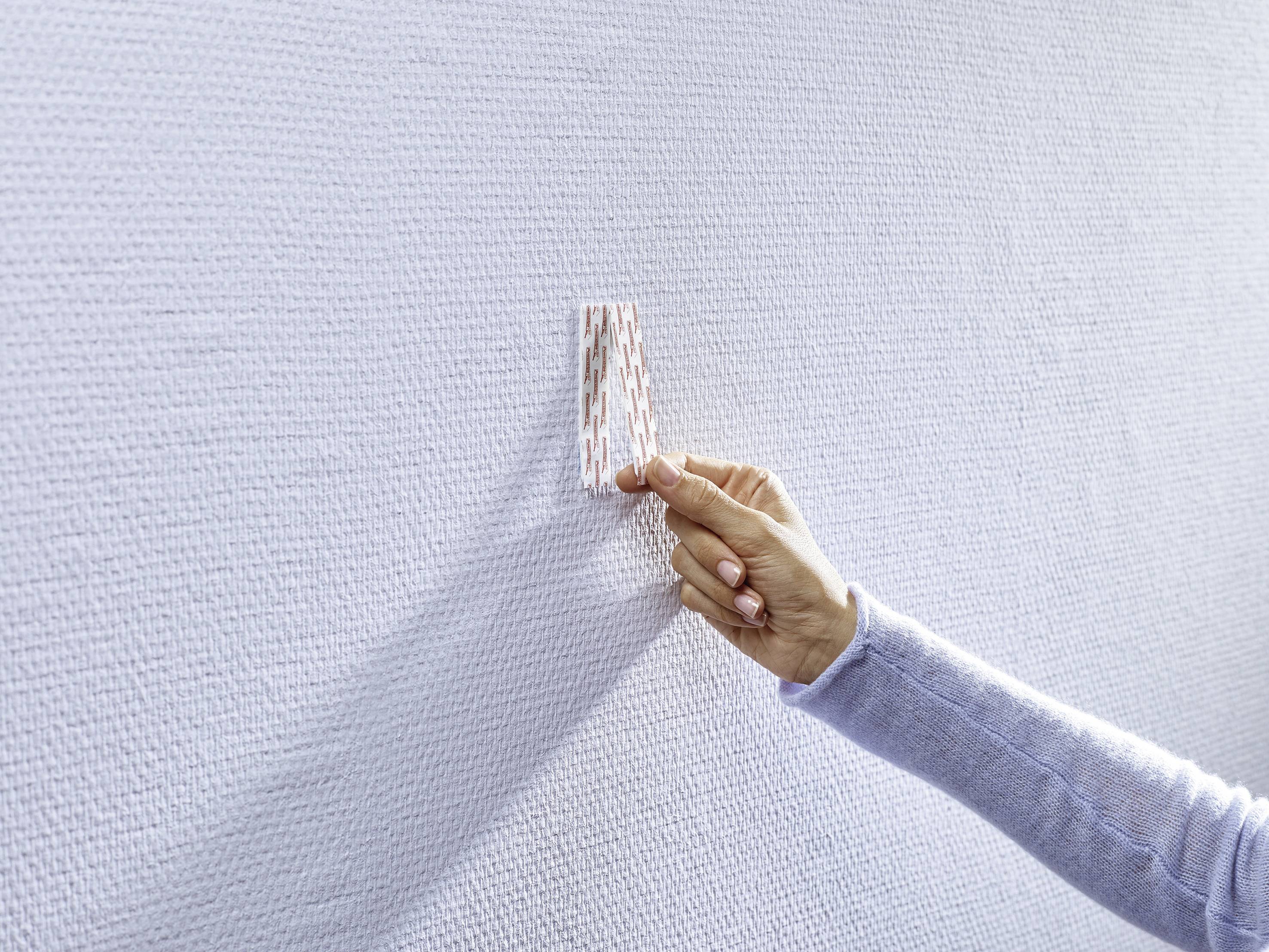 Eine Hand hält eine gestreifte Karte an eine weiße, strukturierte Wand, möglicherweise um Farbe oder Design zu überprüfen.