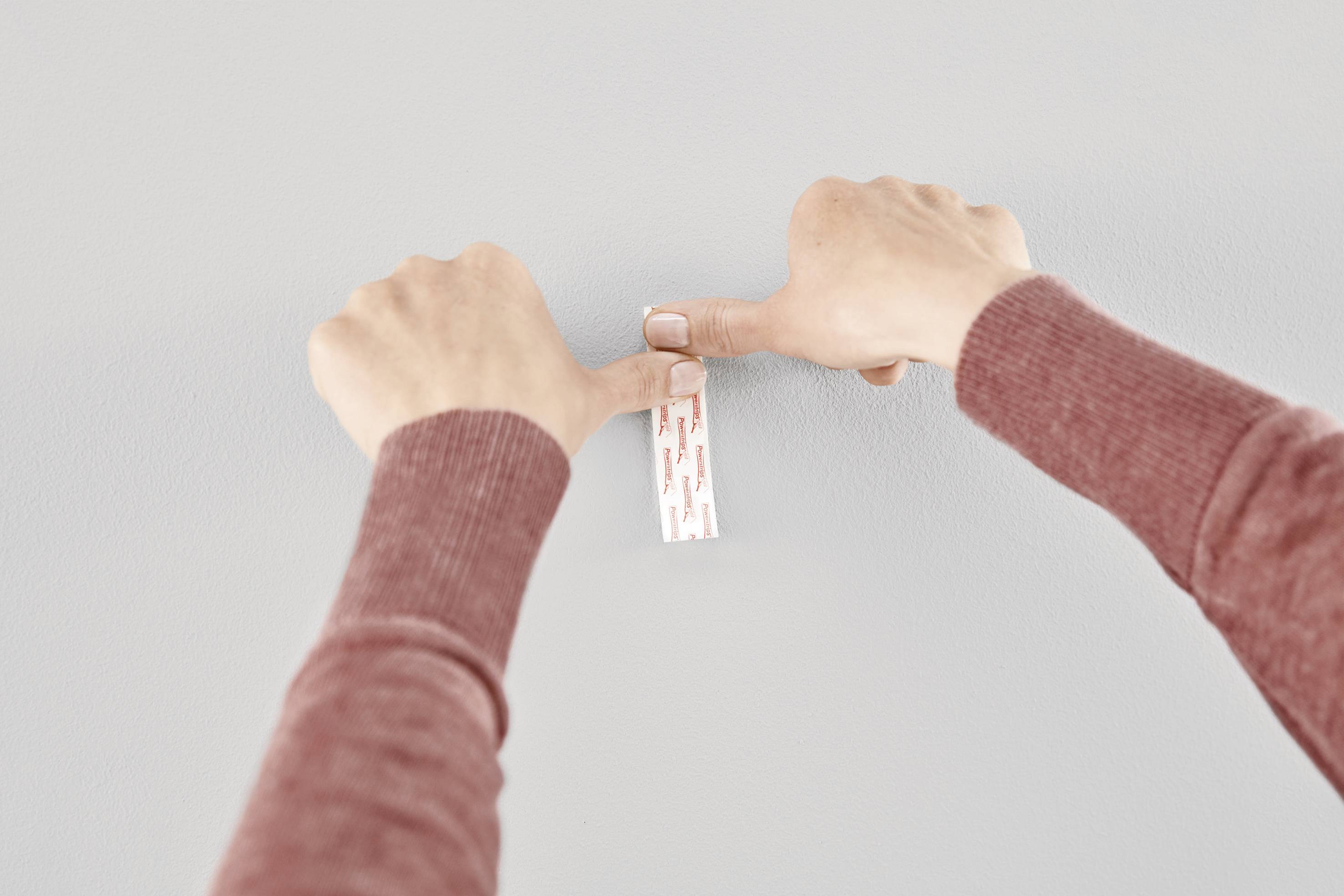 Zwei Hände drücken einen selbstklebenden Streifen an eine Wand, vermutlich um einen Haken zu befestigen.