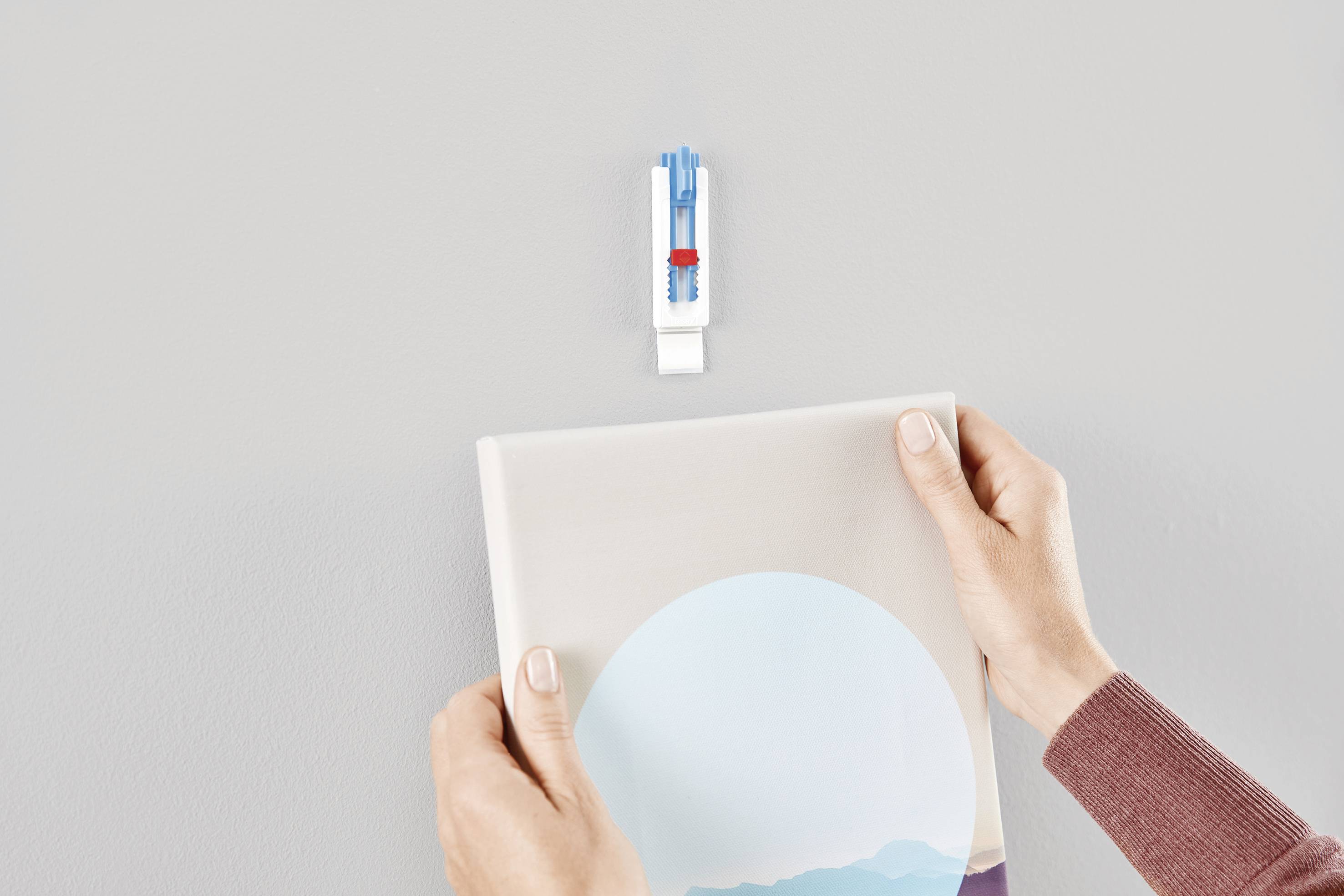 Zwei Hände hängen ein Bild an einen weißen Haken an der Wand auf. Der Haken hat einen blauen Anker und ist vormontiert.