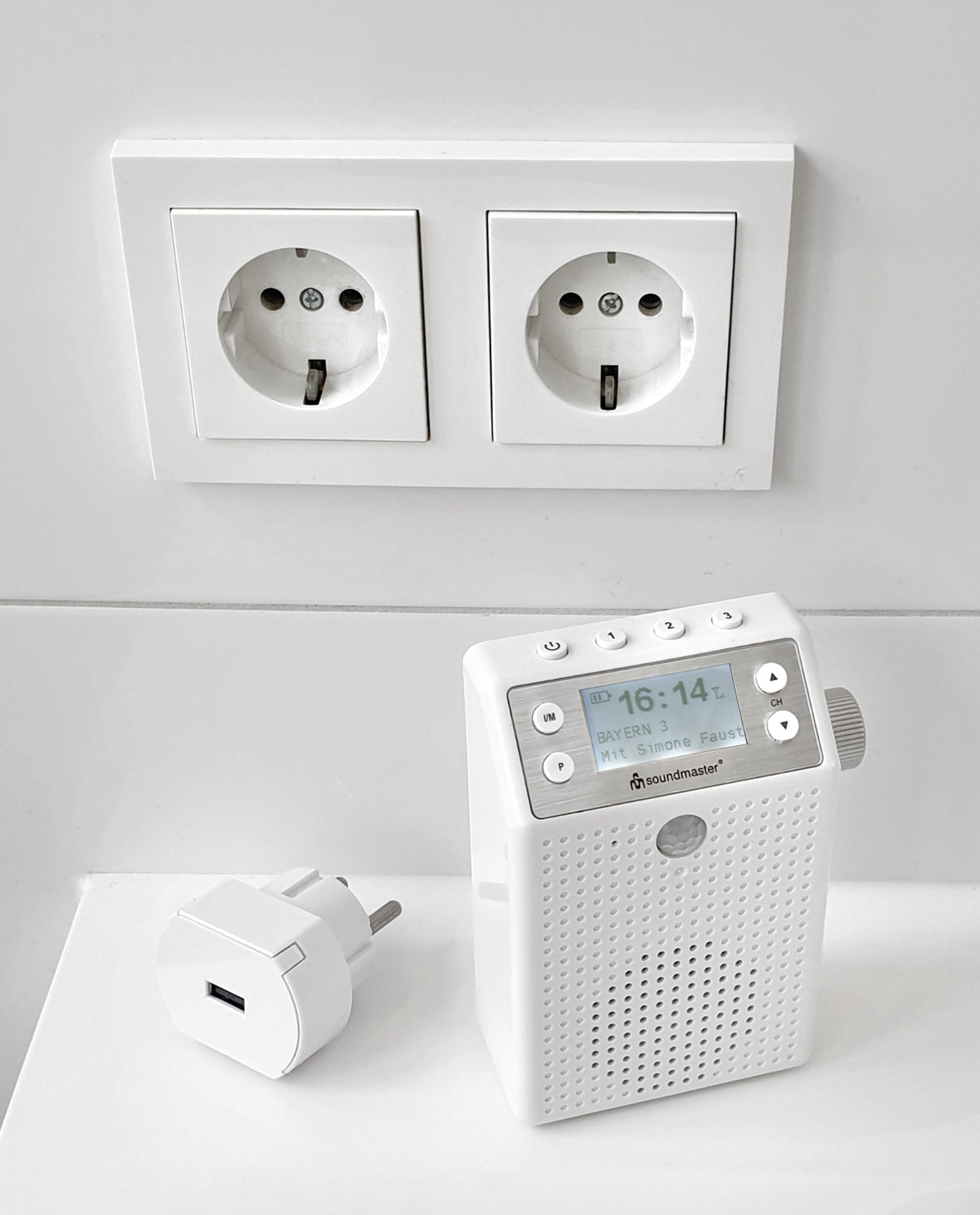Ein weißes UKW-Radio mit digitalem Display zeigt '16:14' an. Daneben liegt ein Adapter vor zwei Doppelsteckdosen an einer weißen Wand.