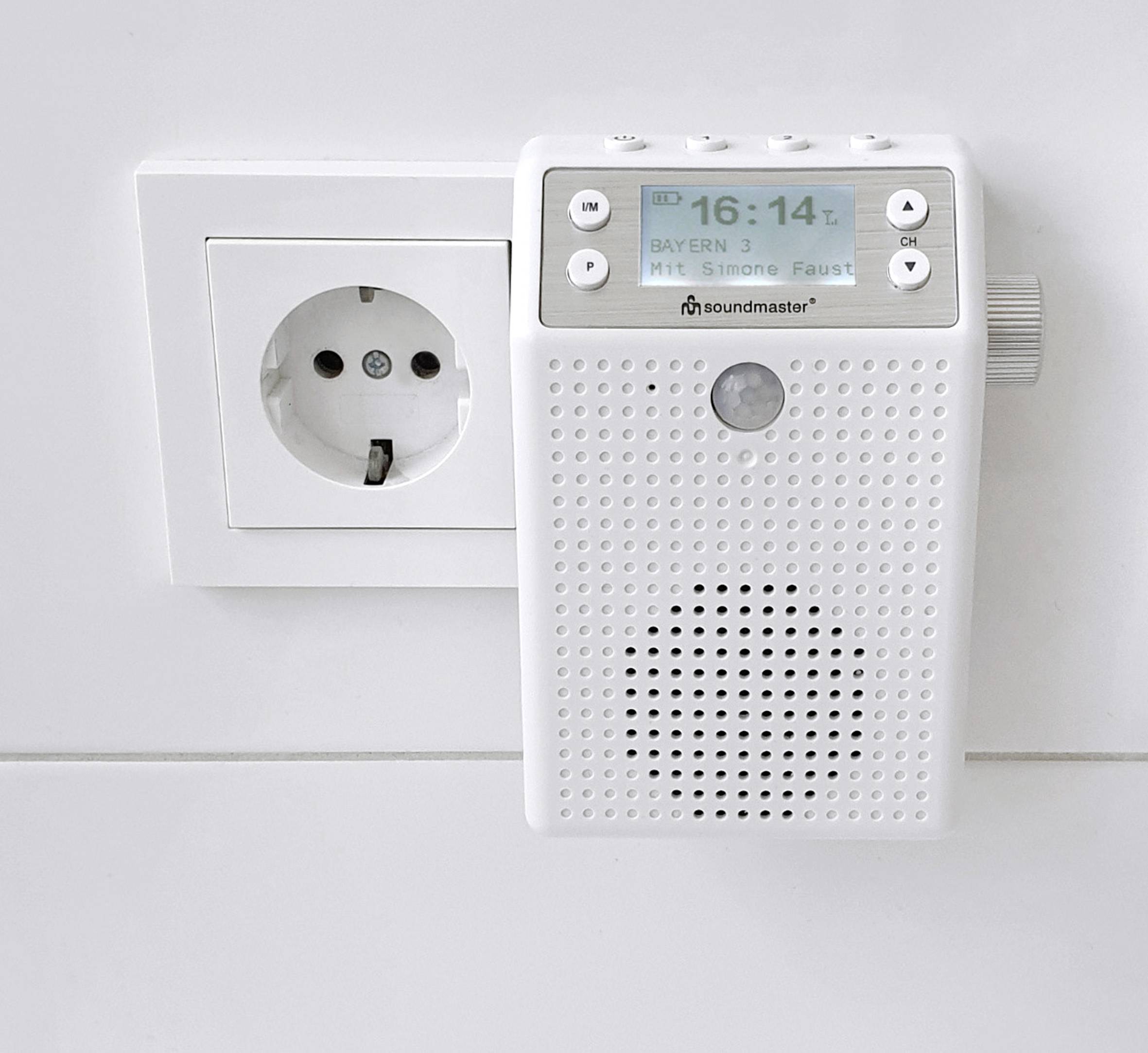 Ein weißes Radio, das an einer Steckdose angeschlossen ist, zeigt die Uhrzeit 16:14 Uhr und den Radiosender Bayern 3 auf seinem Display an.