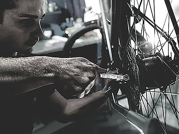 Eine Person repariert ein Fahrrad und konzentriert sich dabei auf das Hinterrad, während sie in einer düsteren Werkstatt einen Schraubenschlüssel verwendet.