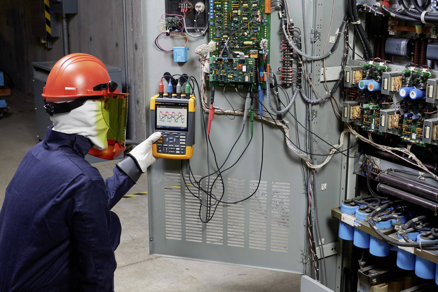 Eine Person in Schutzkleidung misst elektrische Signale an einer offenen Schalttafel mit einem tragbaren Oszilloskop in einer Industrieumgebung.