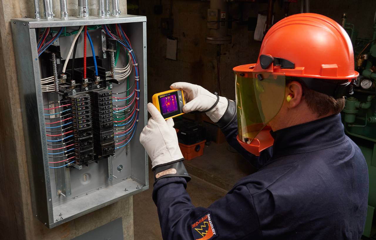 Ein Techniker in Schutzkleidung benutzt eine Wärmebildkamera zur Inspektion eines offenen Sicherungskastens, um elektrische Probleme zu erkennen.