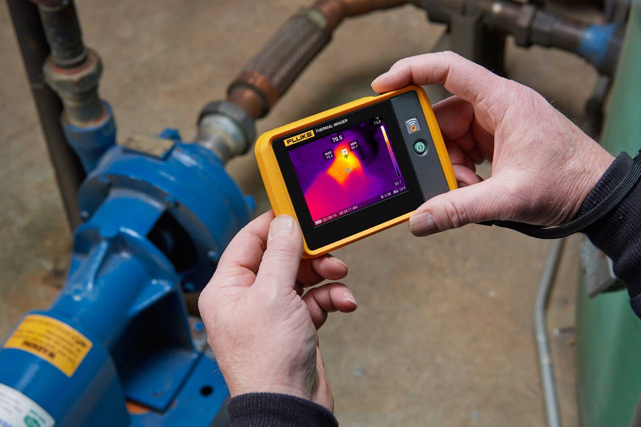 Eine Person hält eine Wärmebildkamera und misst die Temperatur eines blauen Maschinenmotors. Das Display zeigt ein thermisches Bild.