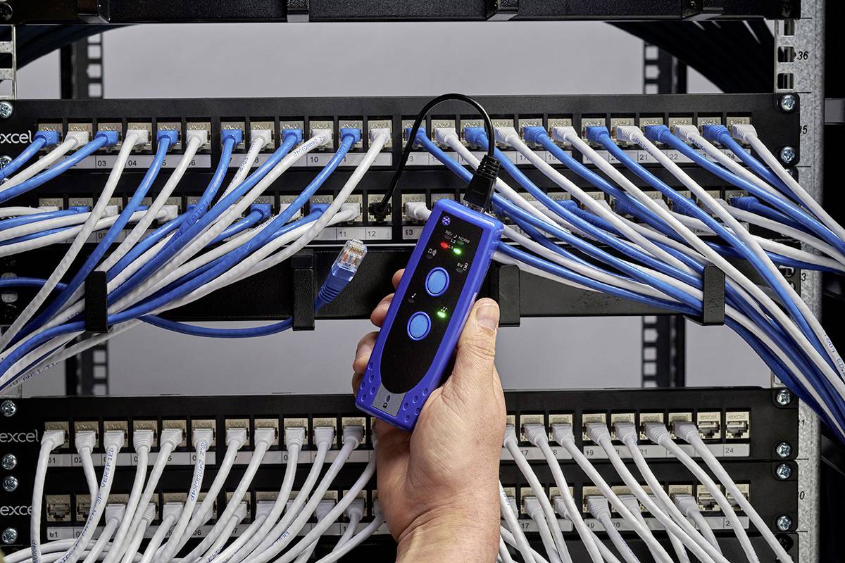 Eine Hand hält ein Netzwerk-Testgerät vor einem Patchpanel, das mit zahlreichen blauen und weißen Ethernet-Kabeln verbunden ist.