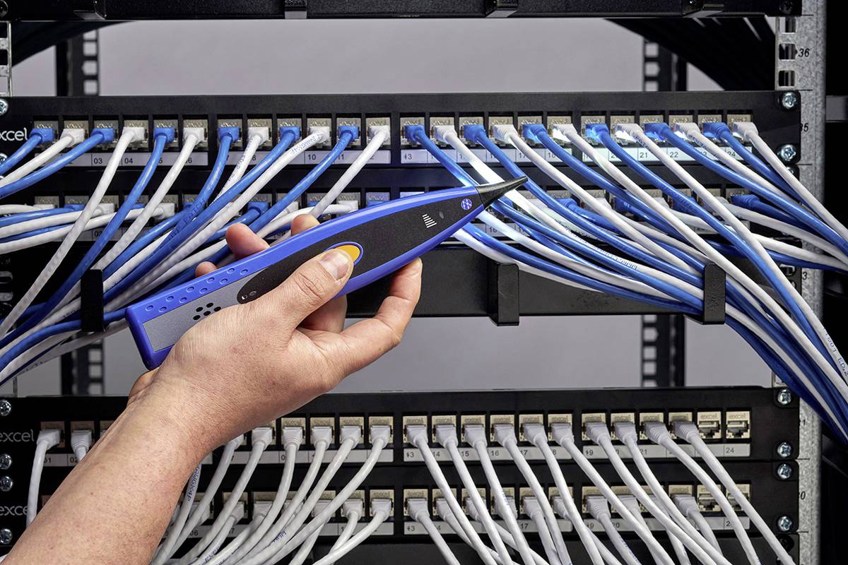 Eine Hand hält ein Prüfgerät, das an Kabel in einem Serverraum angeschlossen ist. Verschiedene Kabel verbinden Netzwerkkomponenten.